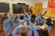 A group of young children in a classroom standing in a circle, wearing matching striped smocks, with their arms raised and hands joined above their heads as part of an activity. The classroom has colorful decorations on the walls, a large blackboard, and a digital screen