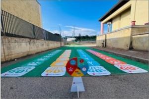 Outdoor game mat with colorful numbered lanes and a spinner in front, placed between two buildings under a blue sky