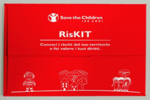 Red box with the Save the Children logo and the title “RisKIT.” The subtitle reads: “Know the risks in your area and claim your rights.” The design includes simple white line drawings of a child, houses, trees, and a group of people, with one speech bubble saying “I think so!”